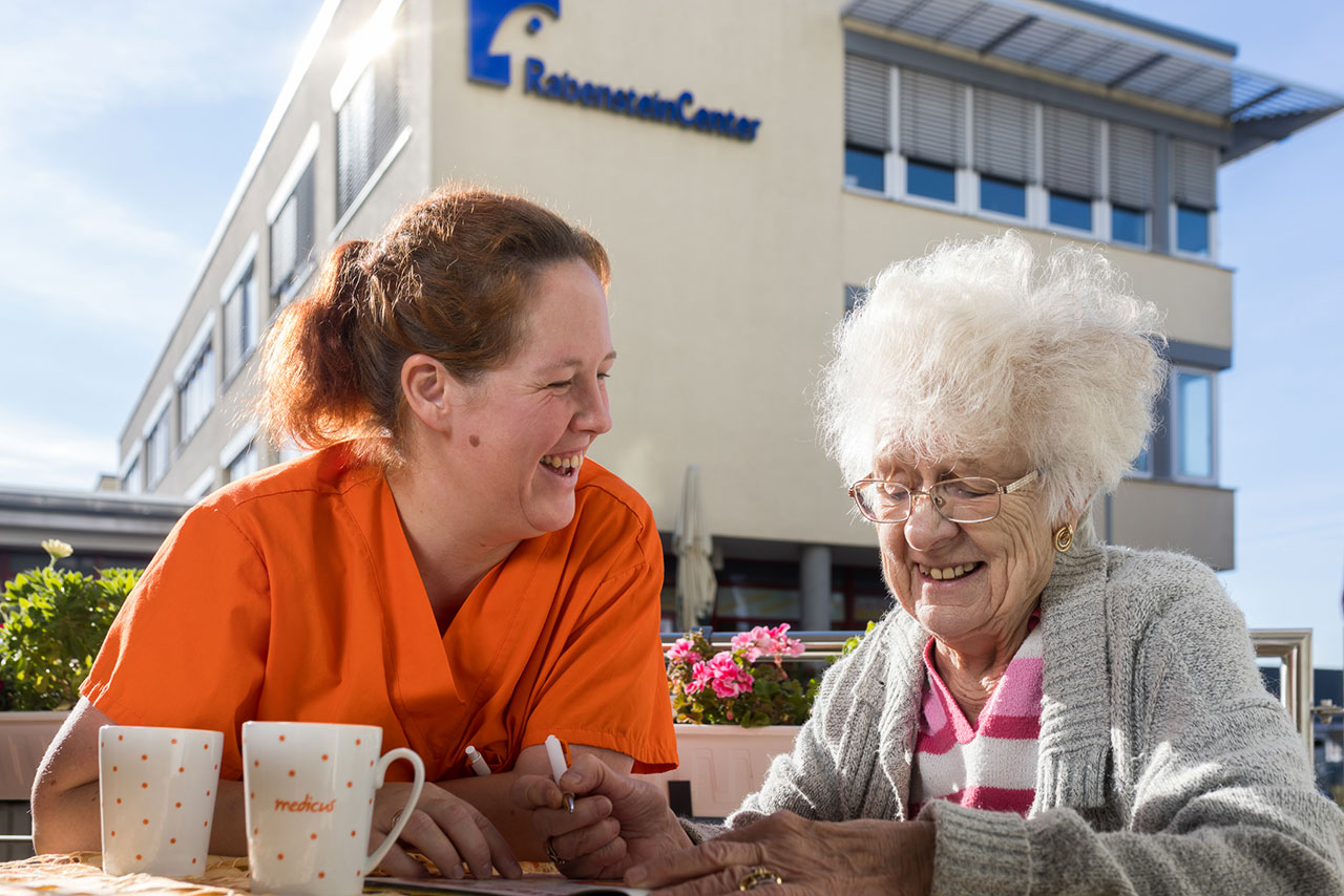 Fröhliche Seniorin und Pflegerin haben Spaß beim Kreuzworträtsel lösen auf der Terrasse