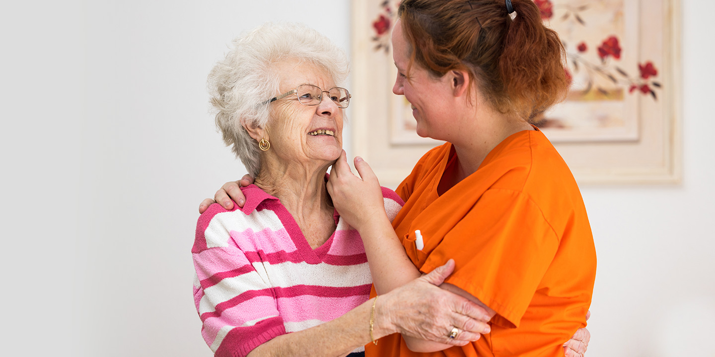 Schwester in orangem Kasack umarmt fröhlich lächelnde Seniorin